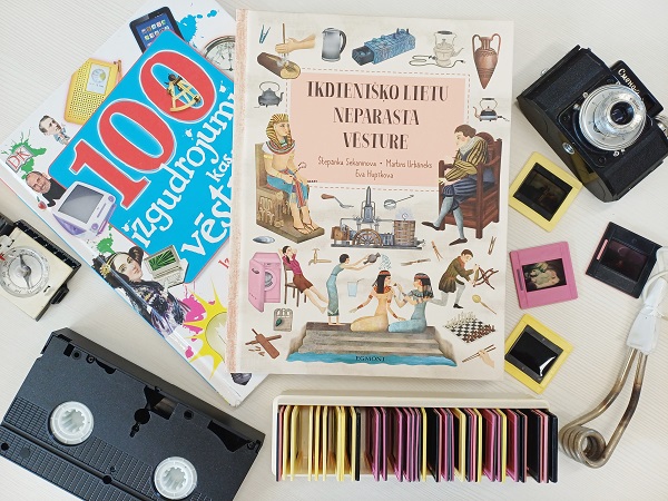 Grāmatas ar nosaukumiem “Ikdienisko Lietu Neparasta Vesture” un “100 Izgudrojumu Kas Izmainija Vesturi” ieskauj vintage priekšmeti, tostarp retro kamera, slaidi un VHS kasete, radot nostalģisku sajūtu.