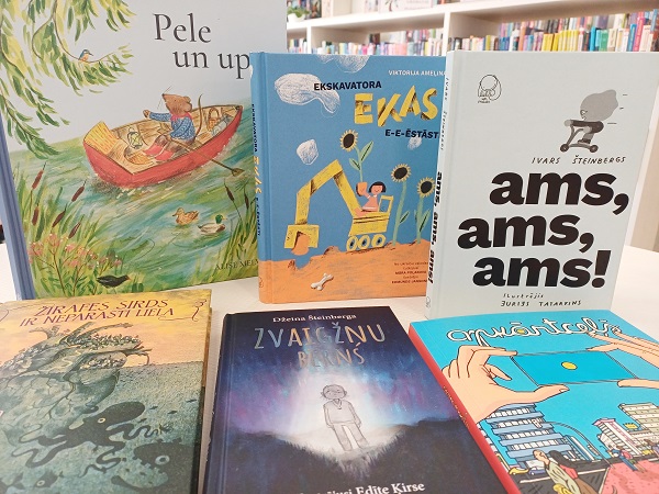 Krāsains sešu bērnu grāmatu klāsts dažādās valodās uz bibliotēkas galda. Uz vākiem ir dzīvnieku, tehnikas un abstraktu dizainu ilustrācijas.