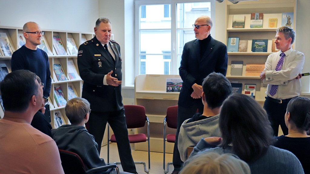 Četru vīriešu grupa stāv telpā, kas piepildīta ar grāmatu plauktiem, uzrunājot auditoriju. Viens vīrietis militārā uniformā runājot žestikulē, paužot autoritāti. Auditorija uzmanīgi klausās. Noskaņojums ir formāls un saistošs.
