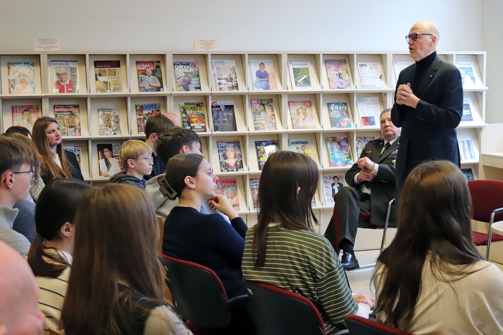 Vīrietis uzvalkā uzrunā uzmanīgu cilvēku grupu telpā, kuras plaukti pilni ar žurnāliem, radot izglītojošu un saistošu atmosfēru.