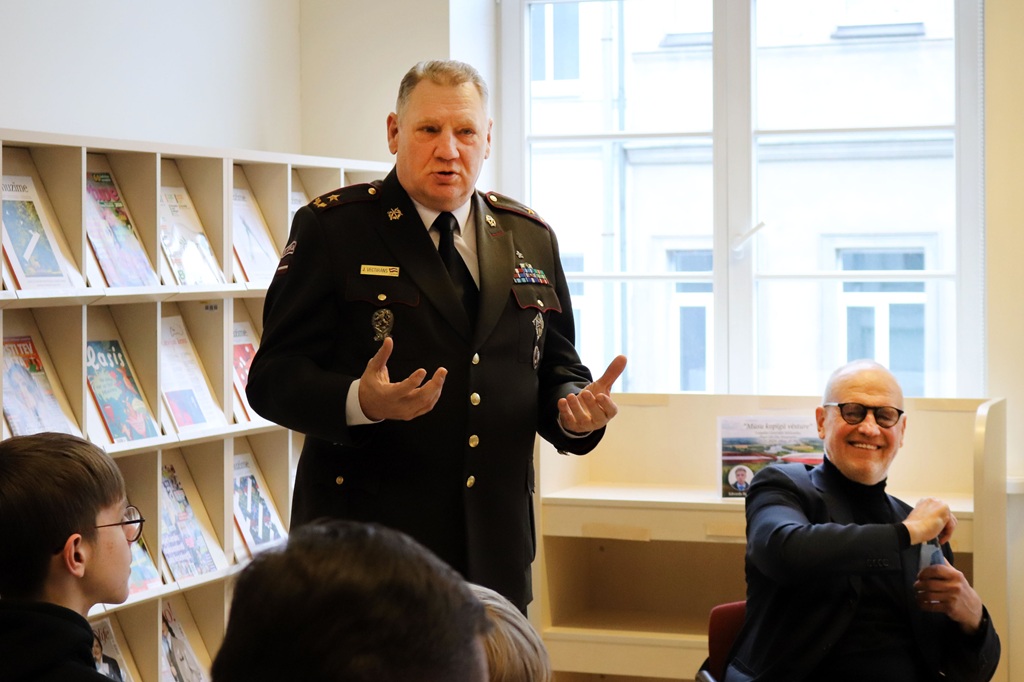 Vīrietis militārā uniformā dzīvīgi uzrunā sēdošu auditoriju telpā ar žurnālu plauktiem; cits vīrietis smaida netālu, radot siltu un saistošu atmosfēru.