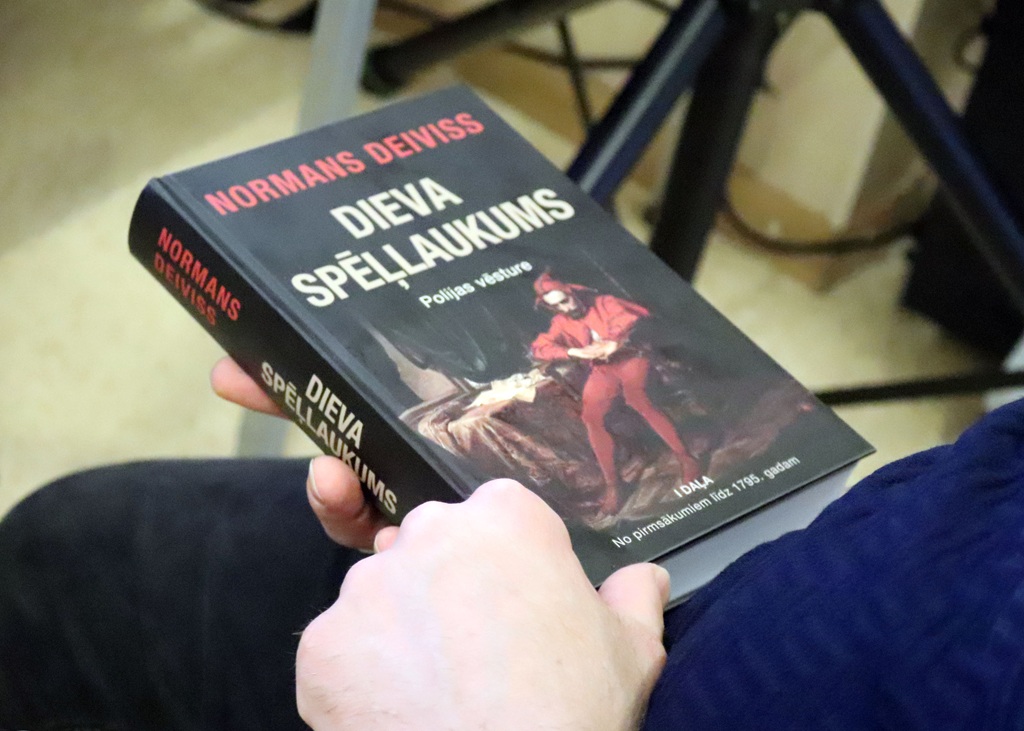 Persona tur rokās Normana Devisa grāmatu ar nosaukumu "Dieva spēle". Uz vāka attēlota sarkanā tērpta figūra, kas lasa dokumentu.