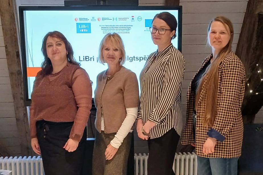 Četras sievietes stāv pie ekrāna, uz kura redzams uzraksts "LiBri". Viņas smaida, radot profesionālu un sadarbības pilnu atmosfēru.