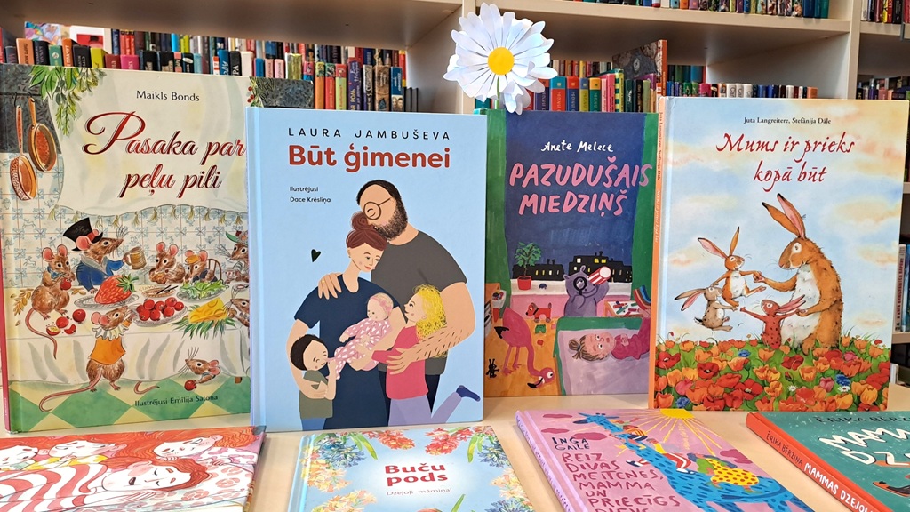 Uz galda ar grāmatu plaukta fonu izvietota krāsainu bērnu grāmatu rinda. Katrai grāmatai ir kaprīzas ilustrācijas un košas krāsas. Kopējais noskaņojums ir dzīvespriecīgs un aicinošs.