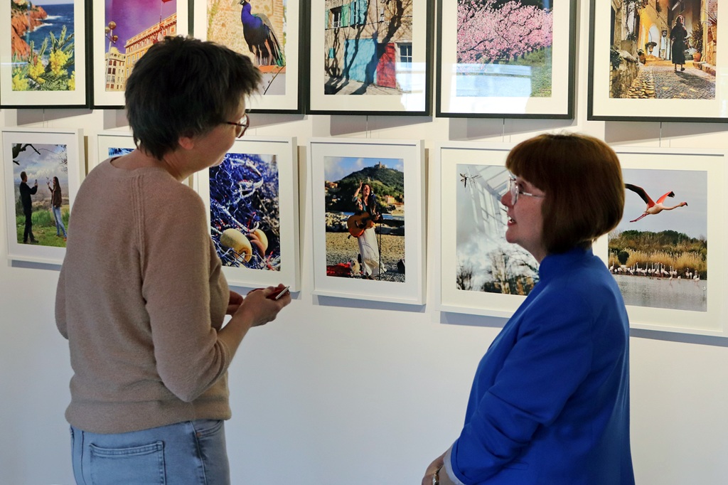 Divi cilvēki iesaistās sarunā, aplūkojot spilgtu fotogaleriju. Fotoattēlos attēlotas dažādas ainavas, dzīvnieki un kultūras ainas, radot dzīvīgu atmosfēru.