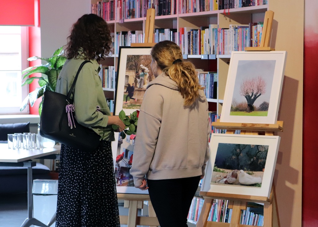 Divas sievietes bibliotēkā aplūko ierāmētas fotogrāfijas uz molbertiem. Fonā plauktos ir grāmatas, radot mājīgu, pārdomu rosinošu atmosfēru.