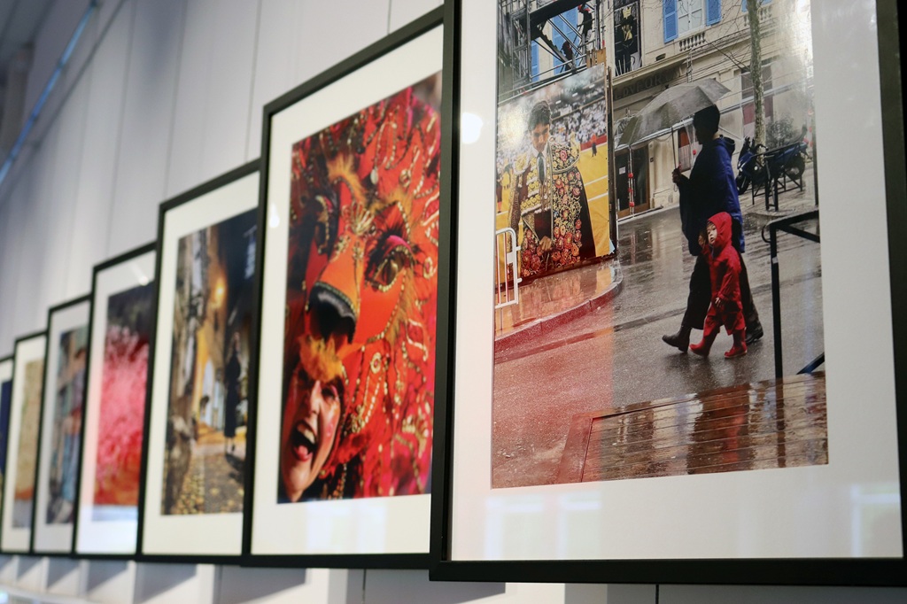 Ierāmētas fotogrāfijas uz galerijas sienas, kurās redzamas dinamiskas kultūras ainas. Spilgts pūķa kostīms un lietaina iela rosina dzīvīgumu un daudzveidību.