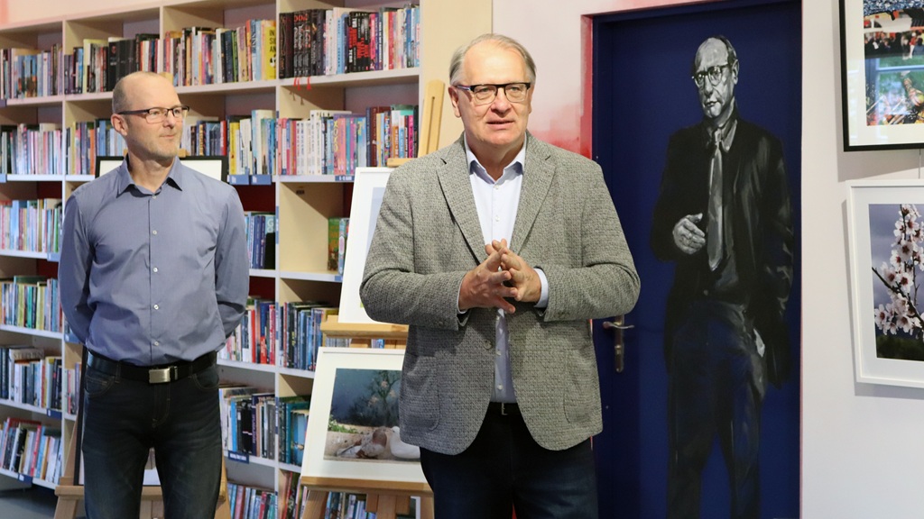 Divi vīrieši stāv bibliotēkā, aiz kuriem atrodas grāmatu plaukti. Viens runā, bet otrs klausās. Izstādīts portrets, kas pauž formālu toni.