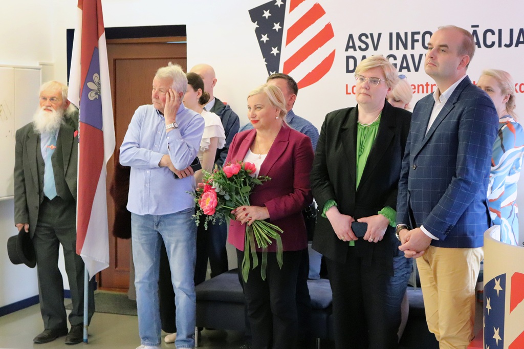 Cilvēku grupa stāv telpā, smaida un pozē. Sieviete bordo krāsas žaketē tur ziedus. Aiz tiem ir zīme ar stilizētu ASV karogu.
