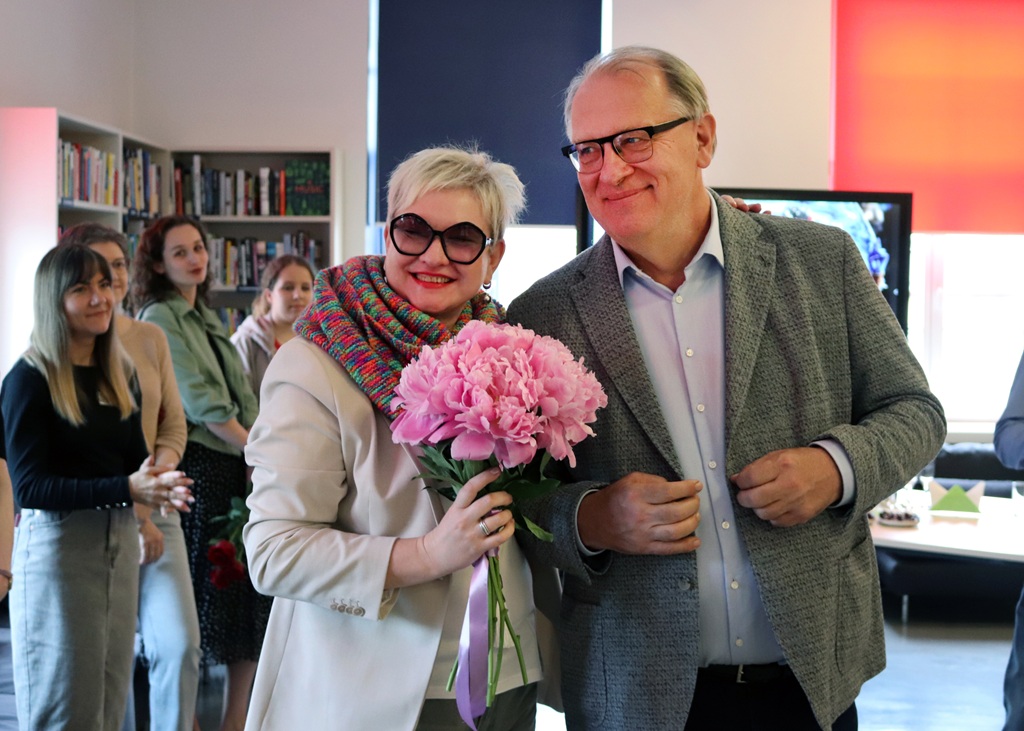 Smaidoša sieviete ar īsiem blondiem matiem un saulesbrillēm tur rokās rozā peonijas blakus vīrietim brillēs mājīgā istabā. Cilvēki fonā laimīgi vēro notiekošo.