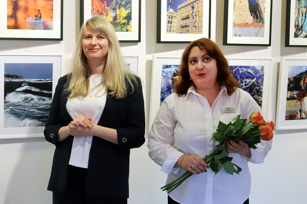 Divas sievietes stāv ierāmētu fotogrāfiju priekšā. Sieviete kreisajā pusē smaida, salikusi rokas, bet sieviete labajā pusē tur oranžu tulpju pušķi.