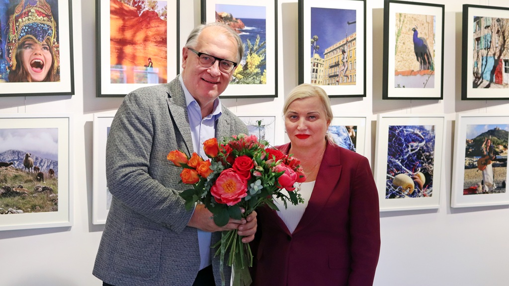 Vīrietis un sieviete smaidoši stāv galerijas sienas priekšā, uz kuras izstādītas krāsainas fotogrāfijas. Vīrietis tur košu sarkanu un oranžu ziedu pušķi.