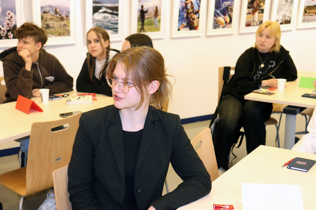 Jauniešu grupa sēž gaišā telpā, iesaistījusies diskusijā. Runā cilvēks melnā uzvalkā, kamēr citi uzmanīgi sēž ap galdiem.