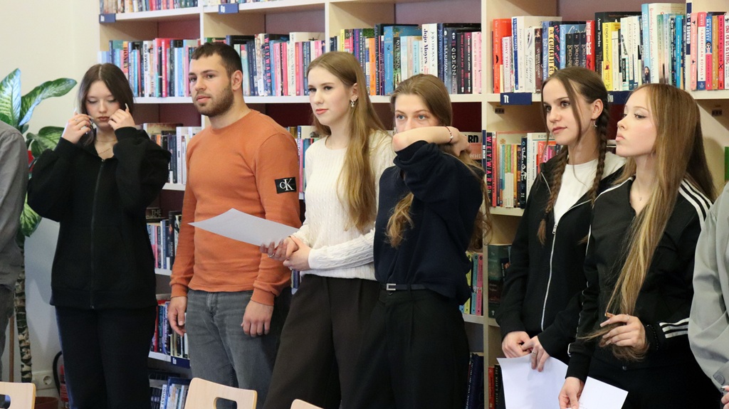 Sešu jauniešu grupa stāv bibliotēkā grāmatu pilnu plauktu priekšā. Viņi šķiet koncentrējušies un uzmanīgi, daži tur rokās papīrus.