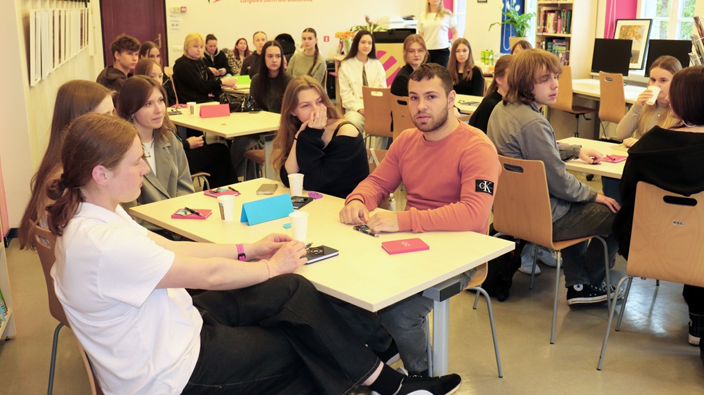 Dažādu skolēnu grupa uzmanīgi sēž ap galdiem klasē, iesaistoties diskusijā. Atmosfēra ir koncentrēta un sadarbīga.