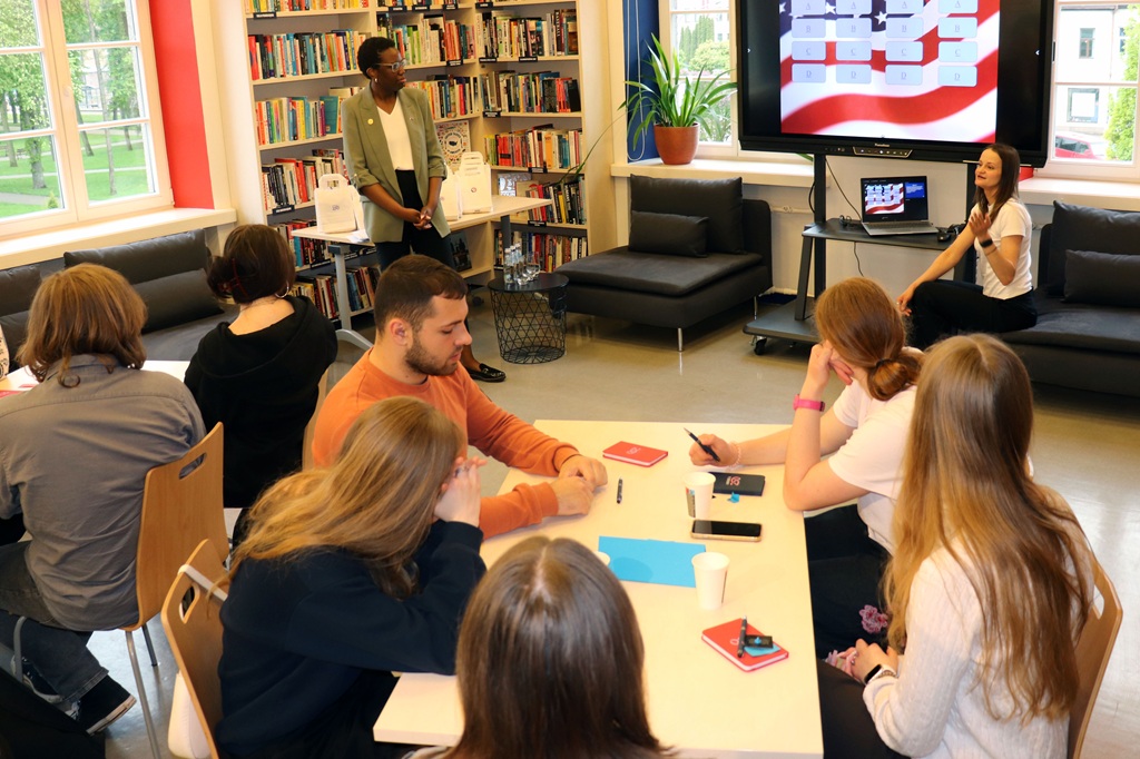 Jauniešu grupa sēž pie galda bibliotēkā un uzmanīgi klausās prezentāciju. Vīrietis stāv pie grāmatu plauktiem, bet sieviete žestikulē pie ekrāna, uz kura redzami slaidi. Atmosfēra ir koncentrēta un izglītojoša.