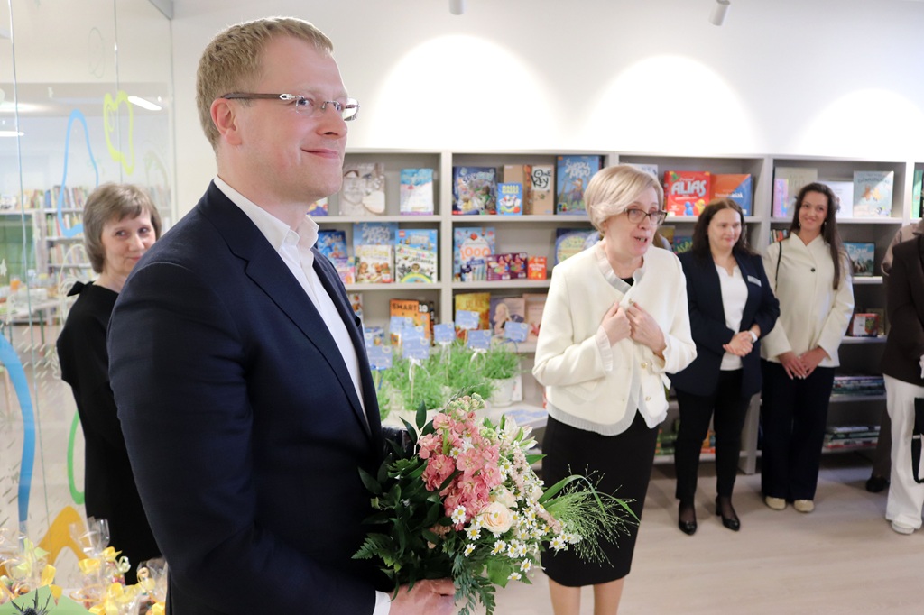 Vīrietis uzvalkā grāmatnīcas vidē smaida un tur rokās pušķi. Blakus viņam dzīvīgi runā sieviete, fonā trīs sieviešu grupa. Atmosfēra ir silta un svinīga.
