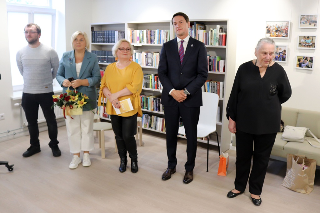Pieci cilvēki stāv bibliotēkā, fonā grāmatu plaukti. Viens tur ziedus. Noskaņojums šķiet formāls un koncentrēts, visi stāv uzmanīgi.