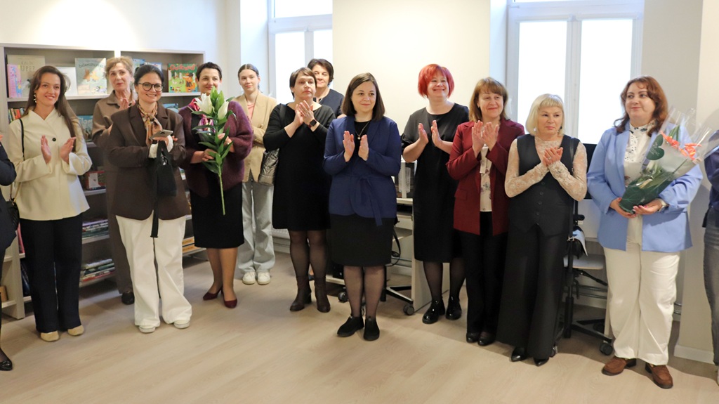Sieviešu grupa stāv telpās, aplaudējot, fonā grāmatu plaukti. Divas sievietes tur rokās pušķus, kas simbolizē svinīgu vai atzinības notikumu.