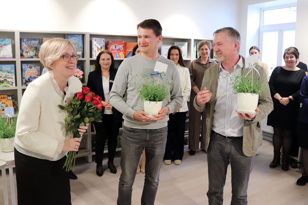 Smaidīgu cilvēku grupa stāv spilgti apgaismotā telpā. Sieviete tur sarkanas rozes, bet divi vīrieši tur podos audzētus augus. Fonā redzami grāmatu plaukti.
