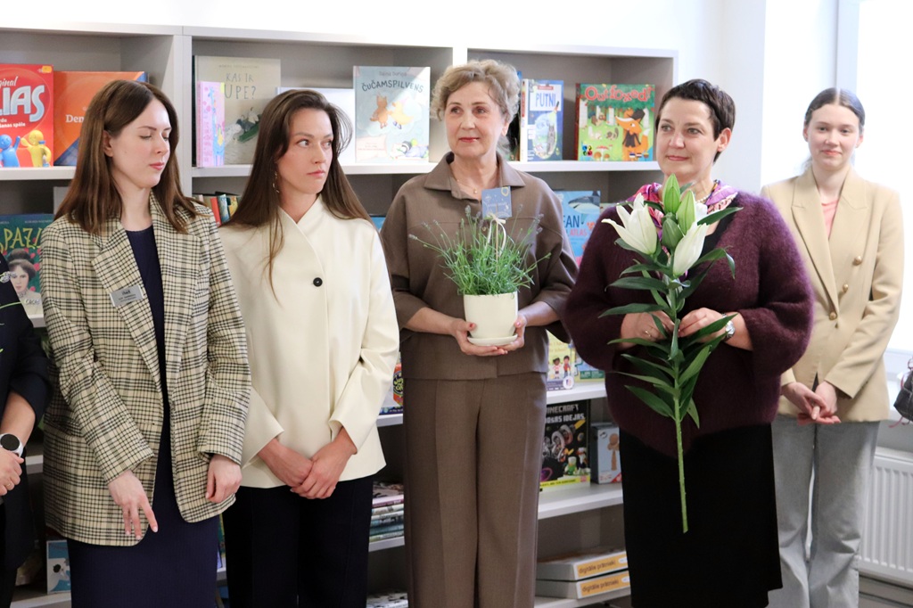 Sešu sieviešu grupa stāv pie grāmatu plaukta, kas pilns ar krāsainām bērnu grāmatām. Divas sievietes tur rokās ziedus, radot siltu un dzīvespriecīgu atmosfēru.