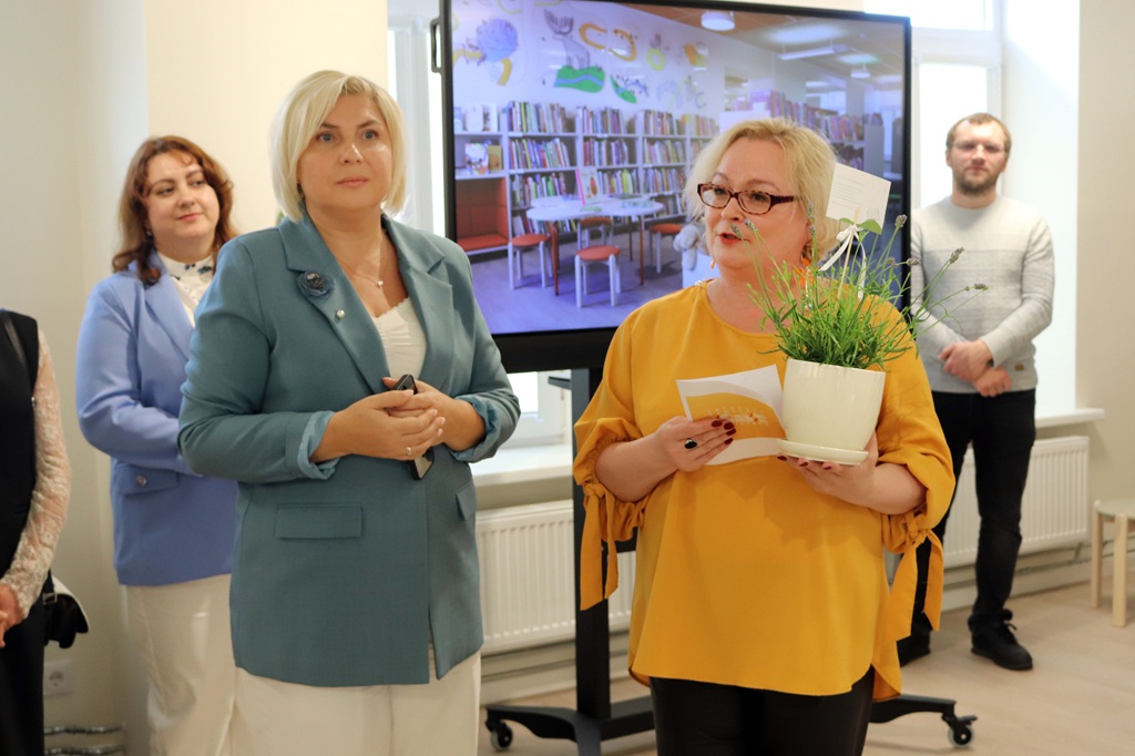 Iekšpusē stāv divi cilvēki, viens zilganzaļā žaketē, otrs dzeltenā blūzē, turot rokās augu. Aiz viņiem uz ekrāna redzama bibliotēka. Atmosfēra ir formāla.