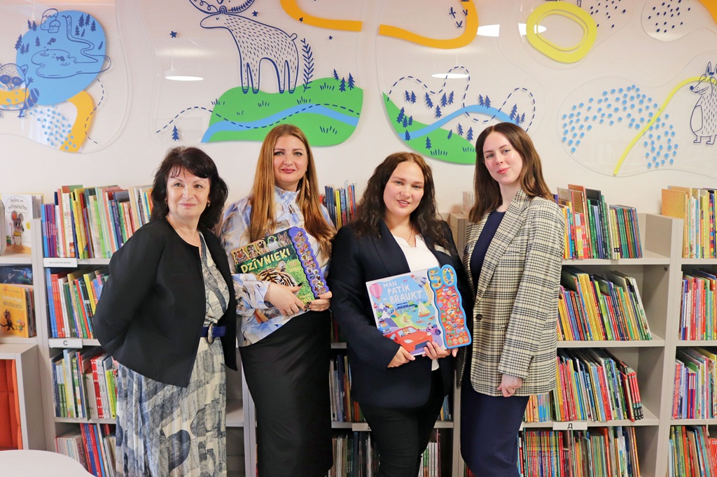 Četras sievietes stāv bibliotēkā, smaida un tur rokās krāsainas bērnu grāmatas. Grāmatu plaukti un rotaļīga sienu māksla rada dzīvīgu atmosfēru.