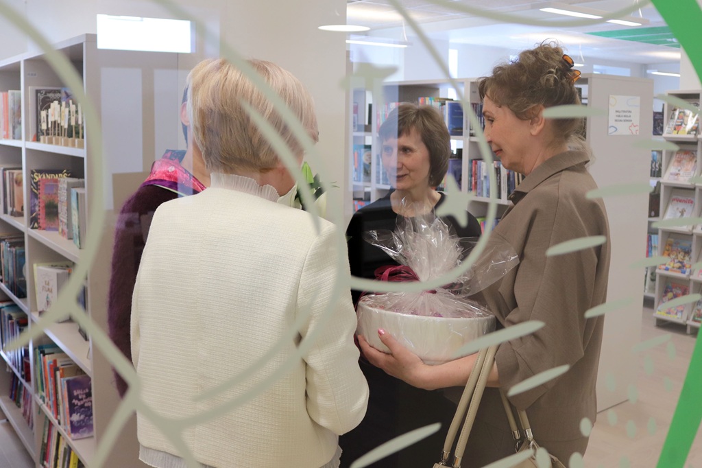 Četri cilvēki sarunājas bibliotēkā, uz viņiem skats caur stiklu ar rakstu. Viens tur dāvanu grozu. Fonā ir grāmatu plaukti.