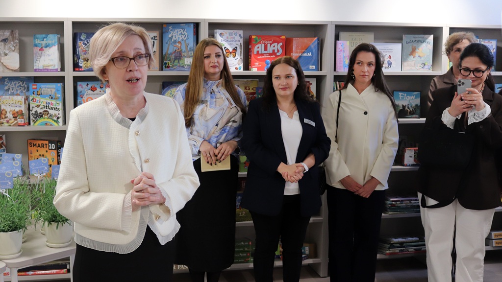 Sieviete runā pie grāmatu plaukta, kas pilns ar krāsainām grāmatām, kamēr citi, oficiālā apģērbā, uzmanīgi klausās. Noskaņojums ir koncentrēts un profesionāls.