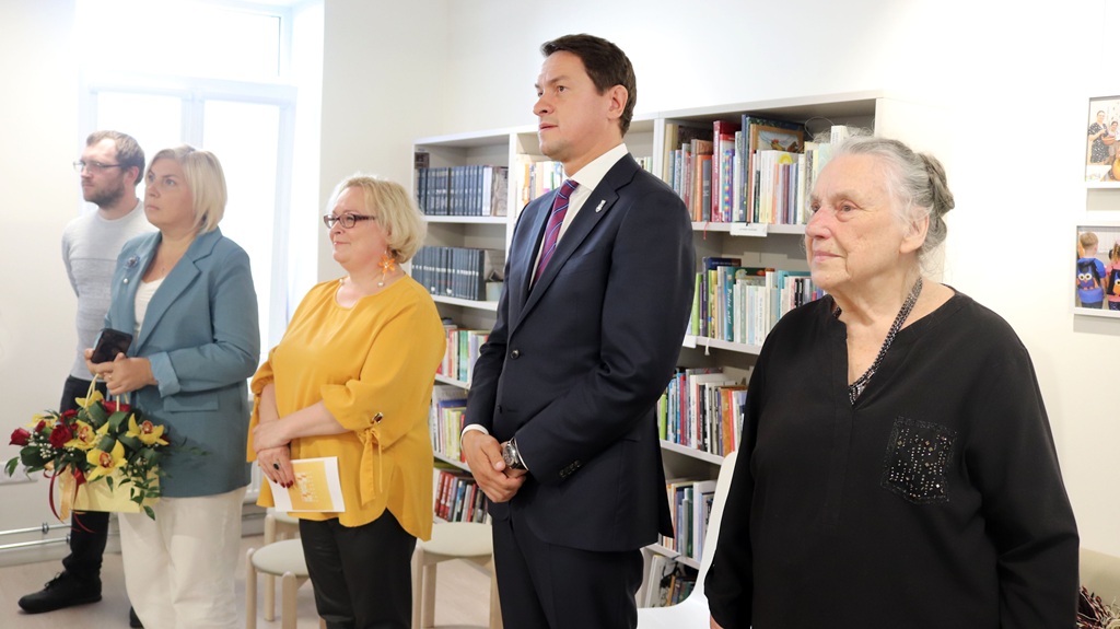 Piecu cilvēku grupa, tostarp trīs sievietes un divi vīrieši, uzmanīgi stāv bibliotēkā. Noskaņojums ir formāls, fonā grāmatu plaukti. Viena sieviete tur rokās pušķi.