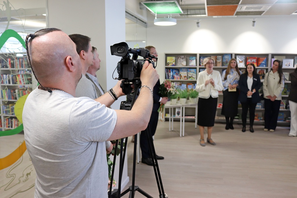 Vīrietis bibliotēkā filmē ar kameru, kas novietota uz statīva, iemūžinot cilvēku grupu, kas uzmanīgi klausās. Plauktos ir grāmatas, radot akadēmisku atmosfēru.