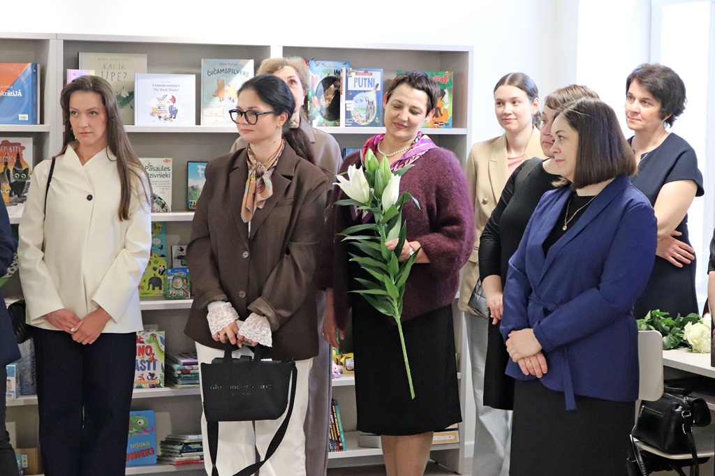 Bibliotēkā stāv sieviešu grupa, sirsnīgi smaidot. Fonā ir plaukti, kas piepildīti ar grāmatām. Viena sieviete tur rokās baltas lilijas, radot svinīgu noskaņu.