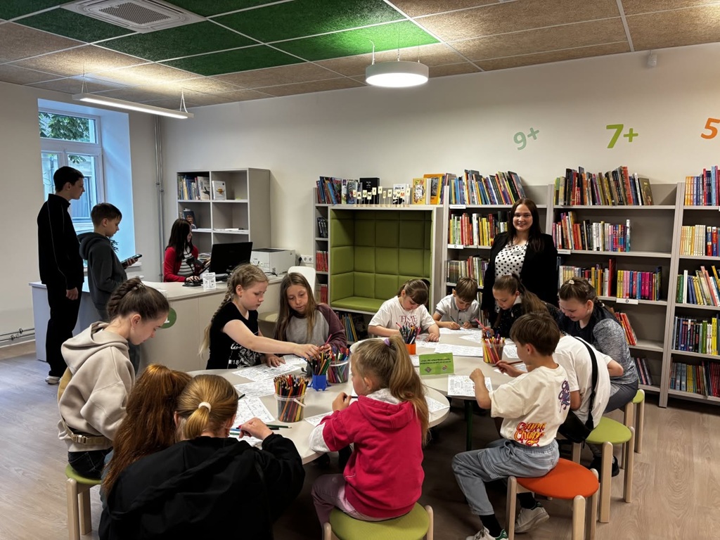 Bērnu grupa sēž ap apaļu galdu gaišā bibliotēkas telpā un radoši darbojas, krāsojot ar zīmuļiem. Fonā redzami grāmatu plaukti un bibliotekāre.