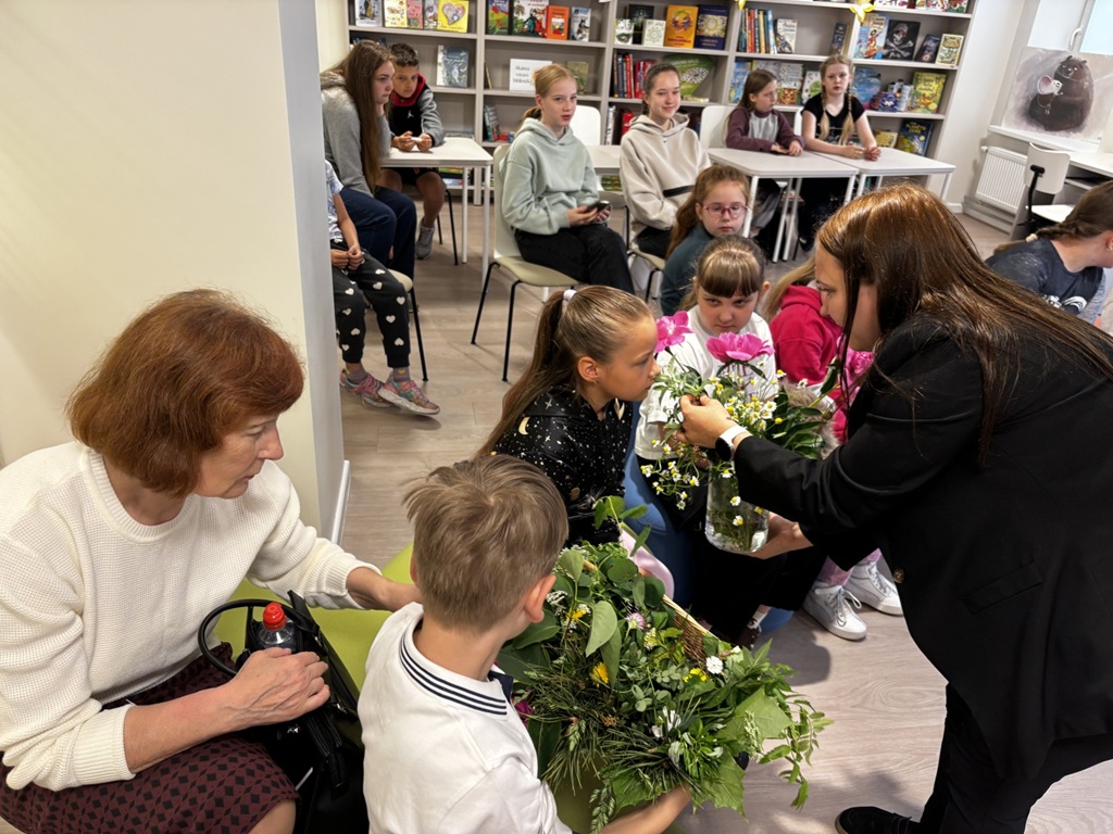 Pasākuma vadītāja tumšā žaketē rāda bērniem stikla vāzi ar pļavas ziediem, ko viena no meitenēm pasmaržo. Priekšplānā zēns tur grozu ar zaļumiem. Aizmugures fonā ir plauks un priekšā tam vairāki bērni, kas sēž uz krēsliem.