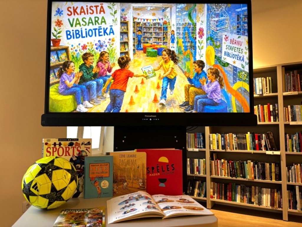 Digitālais displejs rāda bērnus, kas priecīgi spēlējas krāsainā bibliotēkā. Zemāk ir galds ar grāmatām un dzeltenu futbola bumbu. Fonā ir izvietoti grāmatu plaukti.