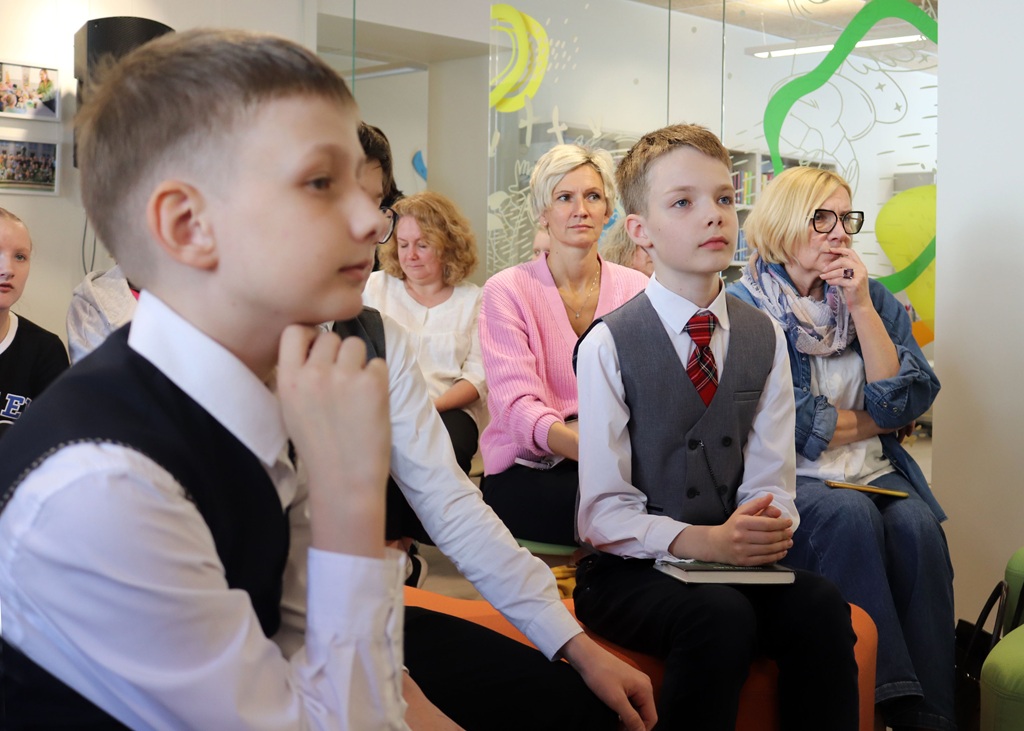 Cilvēki sēž oficiālā gaisotnē, tostarp divi zēni vestēs un kaklasaitēs priekšplānā.