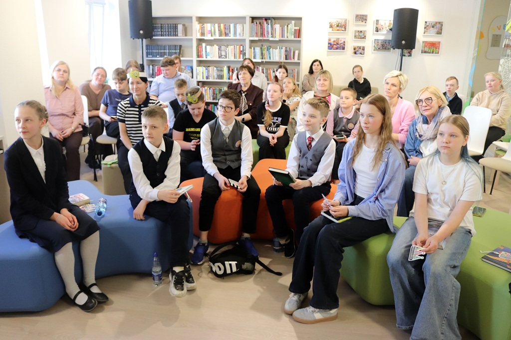 Bērnu un pieaugušo grupa sēž bibliotēkas vidē ar grāmatu plauktiem un ierāmētām fotogrāfijām fonā.