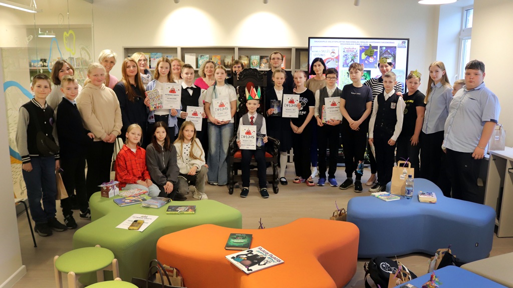 Studentu un pieaugušo grupa stāv kopā bibliotēkā, izliekot apskatei sertifikātus. Telpā ir krāsainas, modernas mēbeles, kas rada svinīgu atmosfēru.