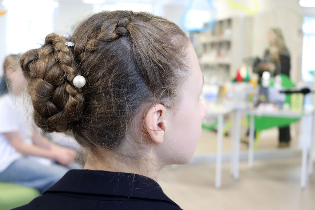 No sāniem redzama meitene ar pītu frizūru, ko rotā neliels pērļu aksesuārs. Viņa sēž gaišā istabā ar izplūdušām figūrām fonā.