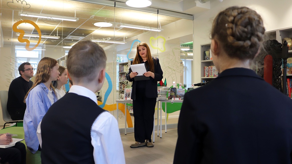Sieviete tumšā uzvalkā uzstājas sēdošai auditorijai modernā telpā ar stikla sienu un krāsainiem dekorācijām.