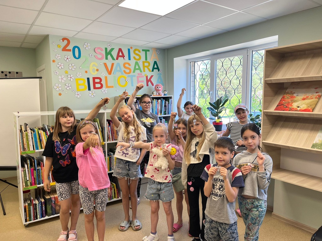 Bērni stāv pie grāmatu plauktiem un priecīgi rāda kamerai savus paštaisītos rokdarbus un piekariņus. Fonā pie sienas krāsains uzraksts '2026 SKAISTĀ VASARA BIBLIOTĒKĀ'