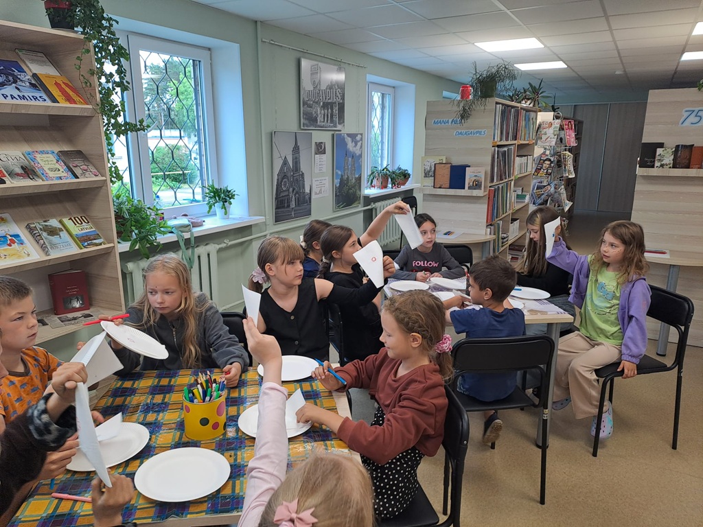 Bērni sēž pie diviem galdiem bibliotēkas telpā, kur radošās nodarbības laikā no papīra šķīvjiem un zīmuļiem veido rokdarbus un rādot tos ir pacēluši uz augšu.