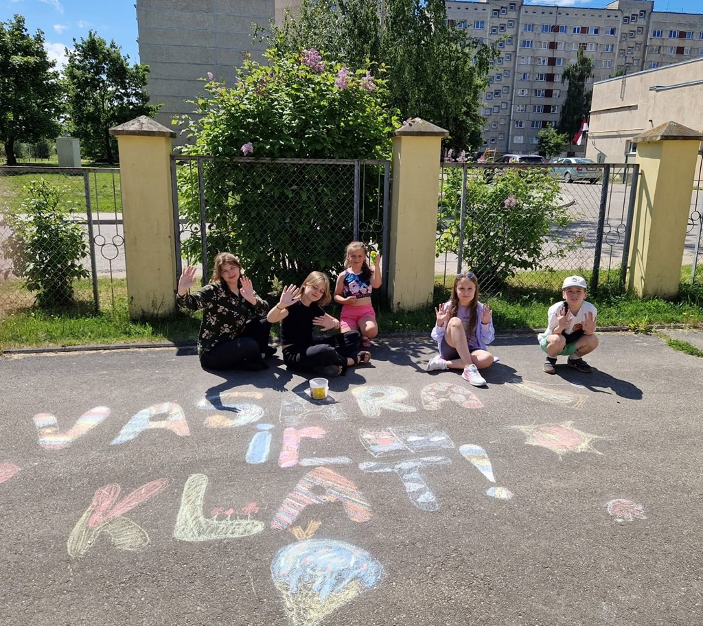 Pieci bērni sēž uz ietves, rakstot un zīmējot ar krāsainiem krītiem, smaida un māj. Fonā redzams žogs un pilsētas ēkas. Jautrs skats.