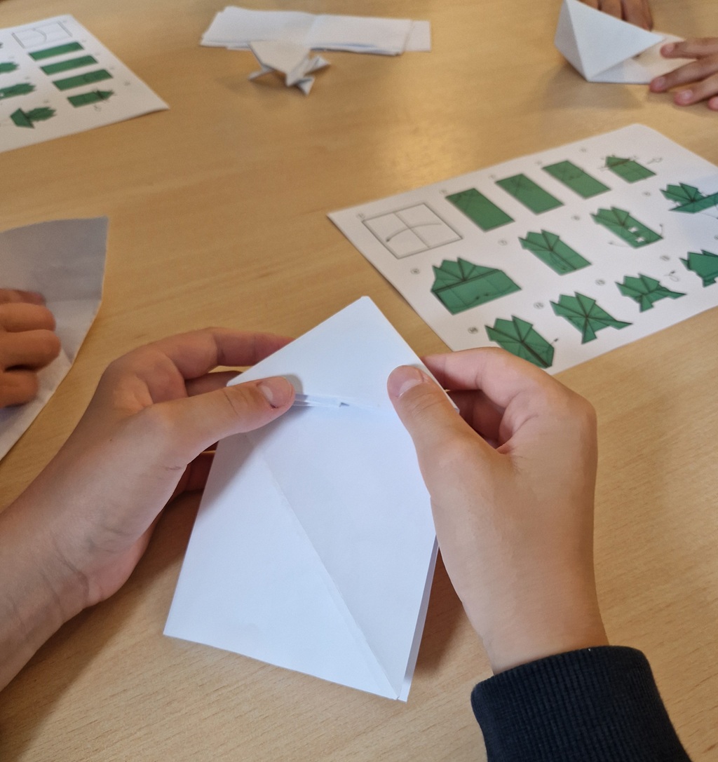 Rokas, kas saloka origami laiviņu no balta papīra. Uz galda ir instrukcijas ar zaļām diagrammām, kā arī citas salocītas papīra formas.