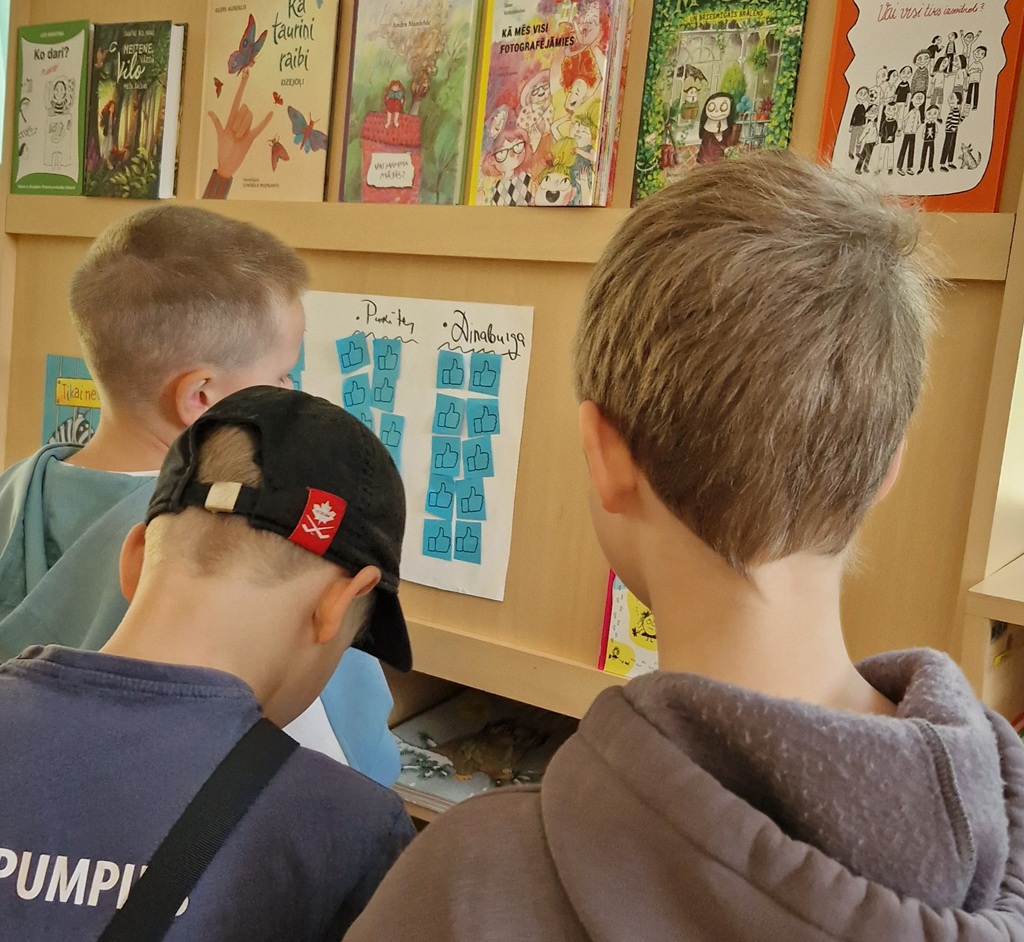 Trīs bērni klases plauktā skatās uz balsošanas zāli ar īkšķiem uz augšu. Aiz tiem ir krāsainas bērnu grāmatas. Aina šķiet interesanta un saistoša.