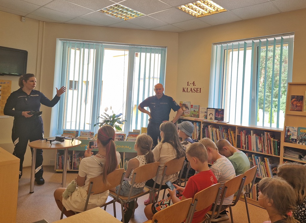 Sieviete un vīrietis, abi formas tērpos, sarunājas ar uzmanīgiem bērniem, kas sēž gaišā bibliotēkā. Viņus ieskauj plaukti ar krāsainām grāmatām un lieli logi.