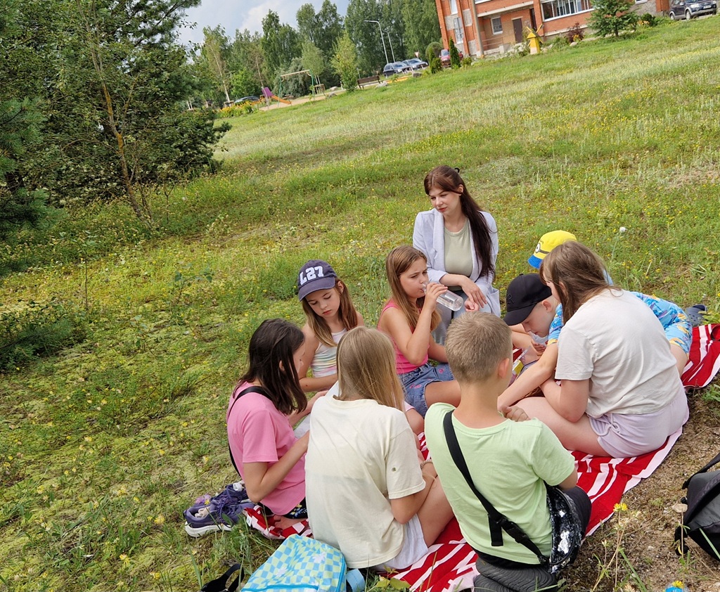 Zāļainā parkā uz sarkansvītrainas segas sēž bērnu grupa, iesaistījušies sarunā. Netālu sēž sieviete, smaidot. Noskaņojums ir mierīgs un dzīvespriecīgs.