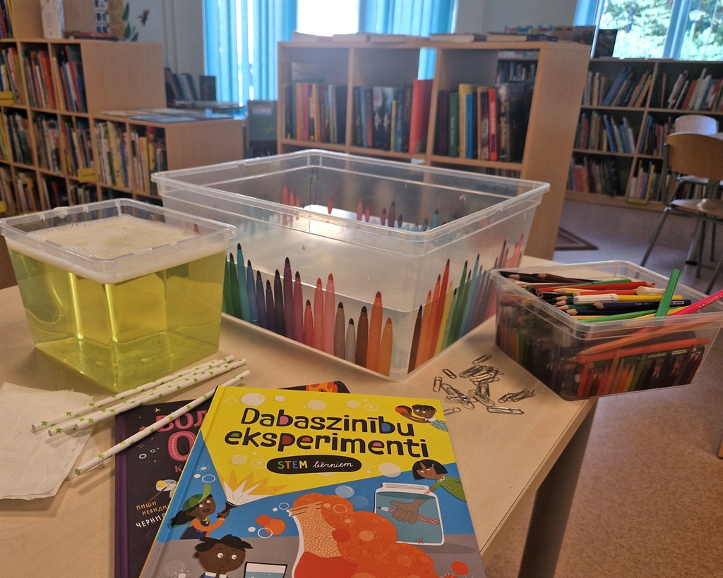STEM mācību iekārtojums gaišā bibliotēkā, kurā ir eksperimentu komplekti ar krāsainiem šķidrumiem, zīmuļi caurspīdīgos traukos un atvērta grāmata par eksperimentiem.