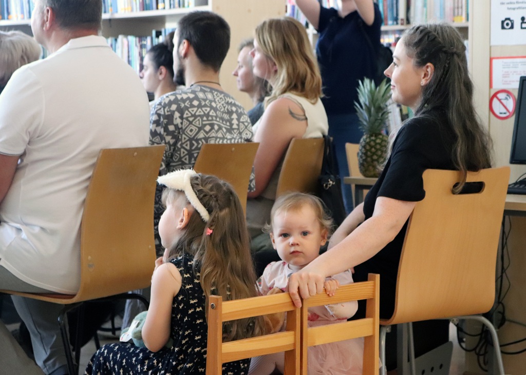 Neliela cilvēku grupa, kurā ietilpst pieaugušie un bērni, sēž bibliotēkā. Bērns ar galvas saiti novēršas, kamēr cits skatās uz kameru. Noskaņojums ir mierīgs.