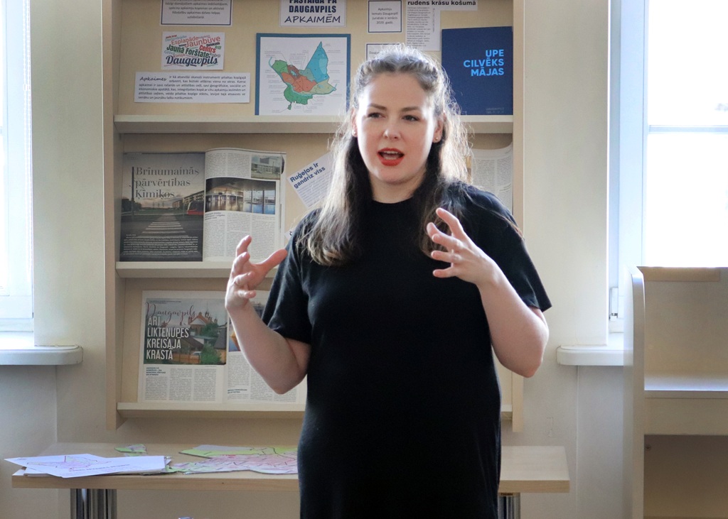 Bibliotēkā stāv cilvēks, kas kaislīgi runā ar izteiksmīgām roku kustībām. Aiz viņiem plauktos ir izvietotas grāmatas un plakāti, kas pastiprina akadēmisko atmosfēru.